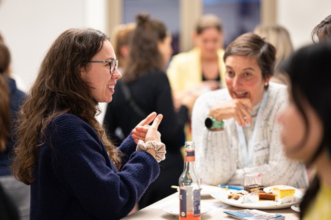 Two women are sitting at a table at the New Year's reception of the Women@DDc network, chatting animatedly and gesturing, while drinks and cake are on the table.