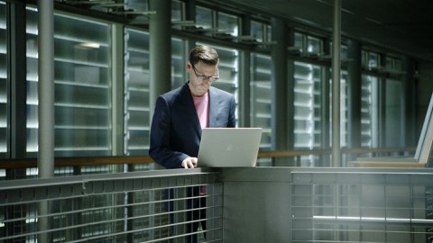  Prof. Koch is in the right part of the photo. He is standing at a railing on which there is an unfolded laptop into which he is looking. In the background, closed external blinds.