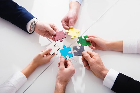 Sechs Hände verschiedener Personen halten sechs verschieden farbige Puzzleteile als Kreis in die Mitte. 