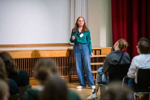 A young woman stands on a stage in a lecture hall, giving a presentation to a seated audience. She is speaking freely and looking toward the audience.