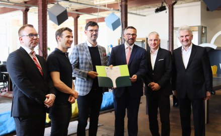 Gruppenbild mit 6 Herren nebeneinander im Anzug. Die zwei Herren in der MIttel halten gemeinsam eine Urkunde in einer Mappe in den Händen. Alle Herren schauen freundlich.