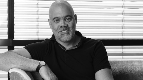 A photo. A black and white portrait of a man sitting on a sofa. In the background, one can see a window with the external blinds.