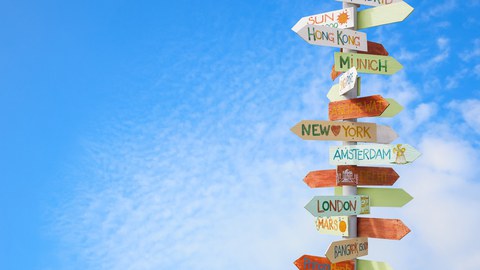 The photo shows a signpost against a blue sky with a few small clouds. There are arrows on the signpost. They bear the names of large cities and point in the direction in which the cities are located.