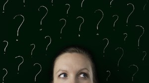 The image shows a photo. In the lower part of the photo you can see the upper half of a face. The eyes are looking to the right. The face is in front of a dark wall. There are many question marks on it.